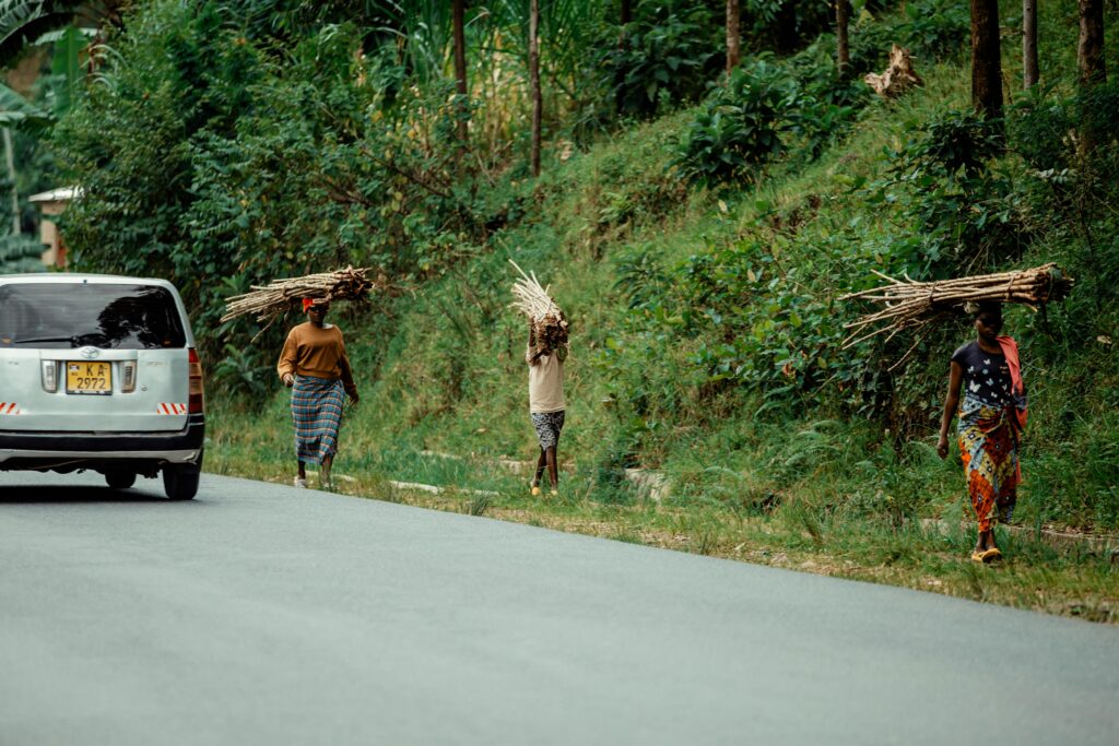 pexels-photo-31448430-31448430 Women walking with firewood on heads by a rural road, next to a passing car.